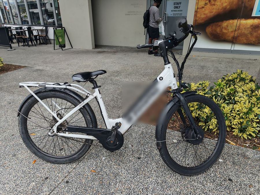 A heavily used, fleet-style delivery e-bike spotted locked up around Brisbane, brand blurred out - dirty frame, no front suspension and a worn, ungreased chain